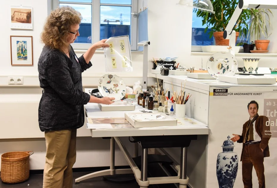 Eine Frau steht in einer Werkstatt und zeigt ein Porzellangefäß.