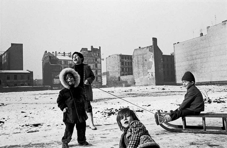 Vier Kinder beim Spiel auf einer leicht mit Schnee bedeckten brachliegenden Fläche, eines der Kinder sitzt auf einem Schlitten, im Hintergrund mehrere verwahrloste alte Mietskasernen ohne Fensterscheiben