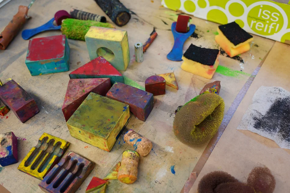 Stempelwerkstatt Auf einem Tisch liegen verschiedene Werkzeuge und Materialien für kreative Arbeiten, darunter bunte Schwämme, bemalte Holzblöcke, Korken und Farbroller.