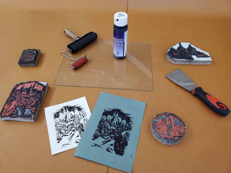 Das Foto zeigt eine Arbeitsfläche, auf der mehrere Utensilien zum Drucken, vier kleine Druckstöcke und zwei Druckgrafiken liegen.