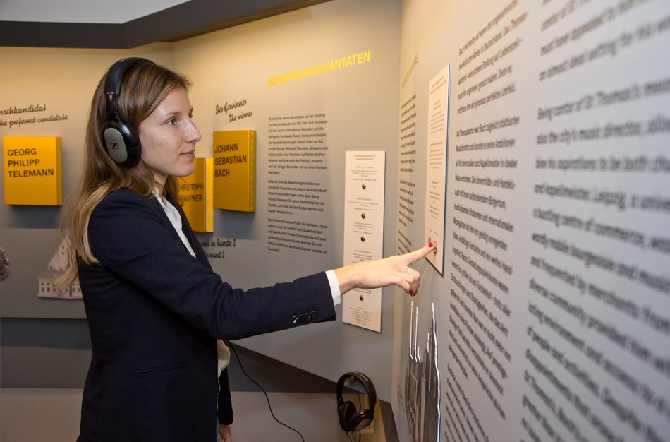 Eine Frau mit Kopfhörern steht an einer mit Ausstellungstexten versehenen didaktischen Wand und drückt auf einen Knopf, um Musikbeispiele aufzurufen.