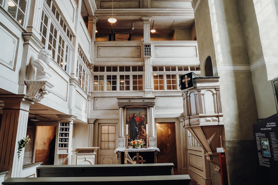 Blick ins Innere einer schnörkellosen Kapelle mit weißen Wandvertäfelungen und drei Emporen. Einziger farbiger Blickfang ist das Altargemälde.