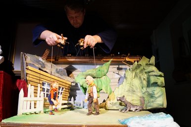 Ein Marionettenspieler führt die Marionetten zweier menschlicher Figuren vor bäuerlicher Kulisse, rechts daneben sitzt lauernd die Marionette eines Wolfes