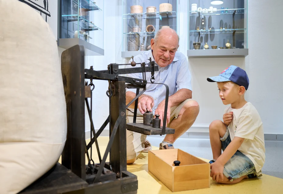 Foto eines älteren Herrn und eines kleinen Jungen, die vor einer historischen Waage hocken und sich am Wiegen eines gefüllten weißen Sacks versuchen