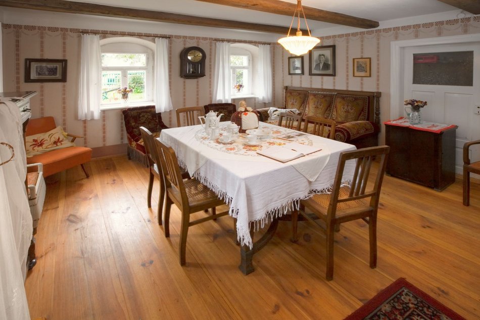Zu sehen ist die Inszenierung einer Wohnstube im alten Bauernhof, mit bedruckter Tapete, Wand-Pendel-Uhr, und einigen historischen Familienfotos an den Wänden. Mittig steht ein großer Esstisch mit weißer Tischdecke und sechs Stühlen.