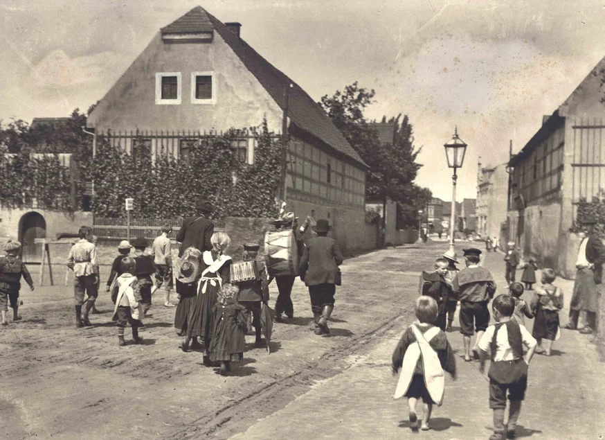 Das historische Schwarzweiß-Foto zeigt, wie eine größere Gruppe Kinder und Erwachsener eine von Fachwerkhäusern gesäumte Straße entlanggehen.