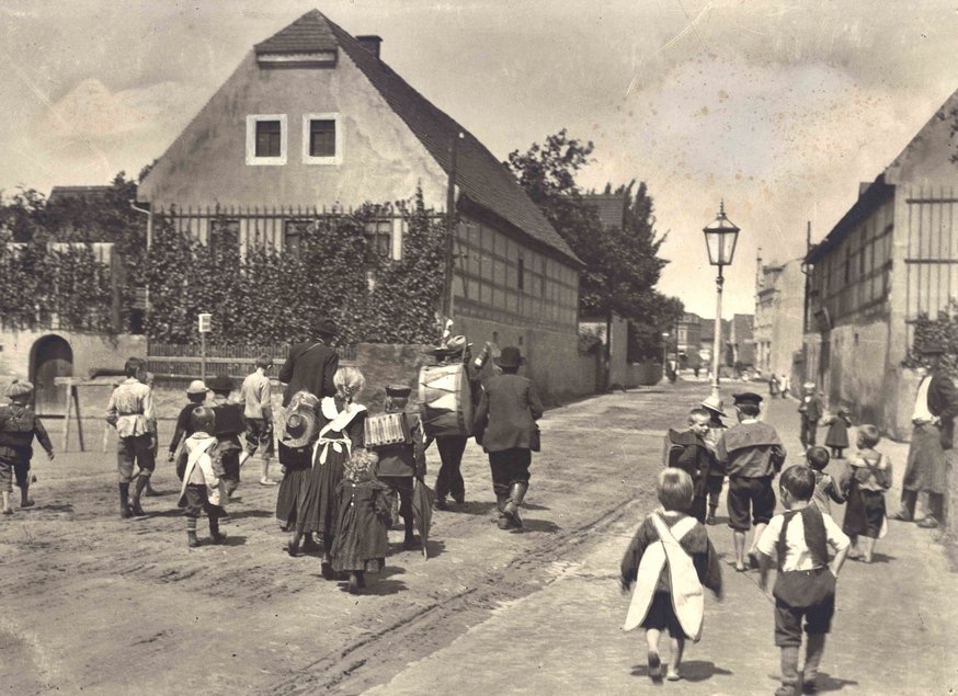Das Schwarzweiß-Foto zeigt, wie eine größere Gruppe Kinder und Erwachsener eine von Fachwerkhäusern gesäumte Straße entlanggehen.