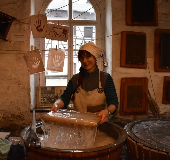 Vorführung Papierschöpfen Eine Frau in historisch anmutendem Kostüm hält über einem Bottich einen Rahmen zum Papierschöpfen