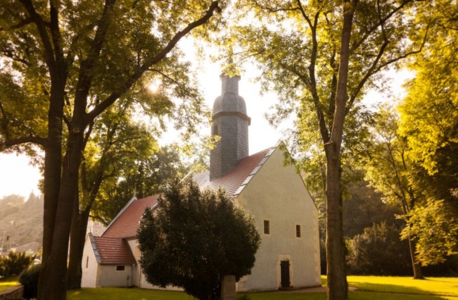 Eine kleine weiße Kirche mit rotem Ziegeldach und schiefergedecktem Turm steht inmitten einer Parkanlage.