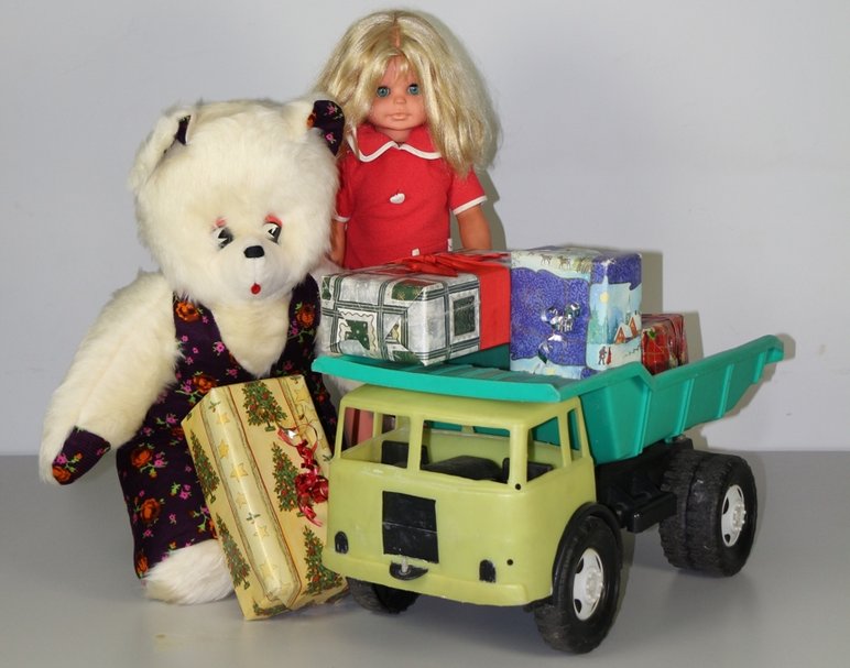 Das Bild zeigt einen Teddy, eine Puppe und einen Kipper sowie weihnachtlich verpackte Geschenke.