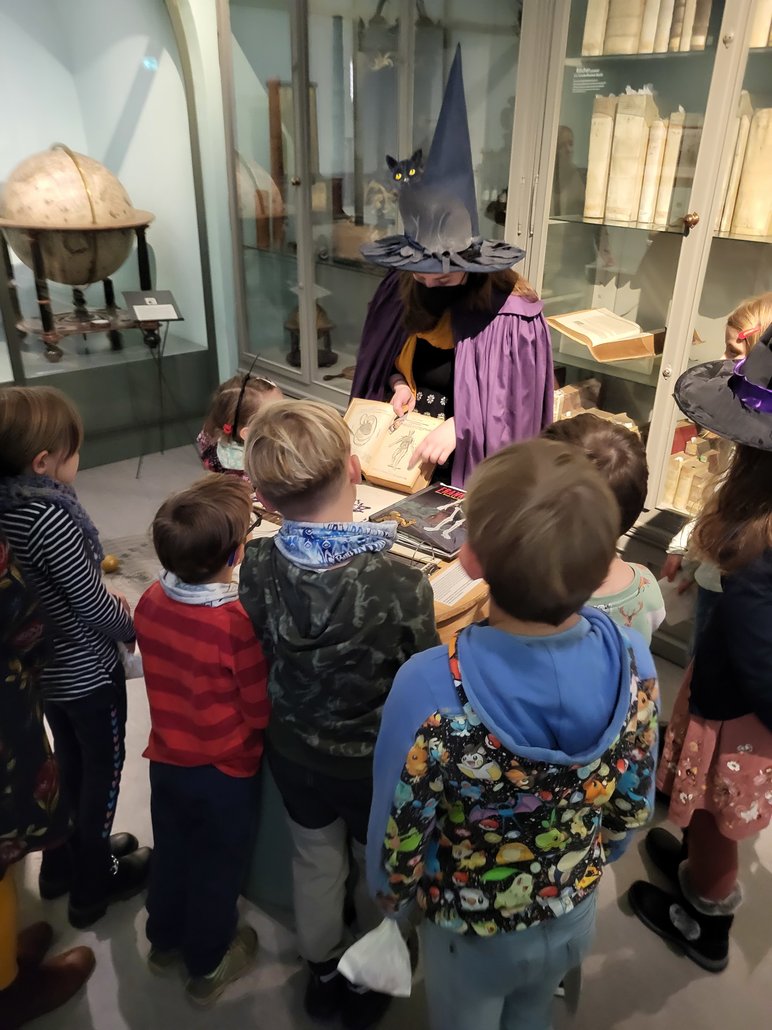 Eine als Hexe verkleidete Frau, die in einem Museumsraum Kindern ein Buch zeigt.