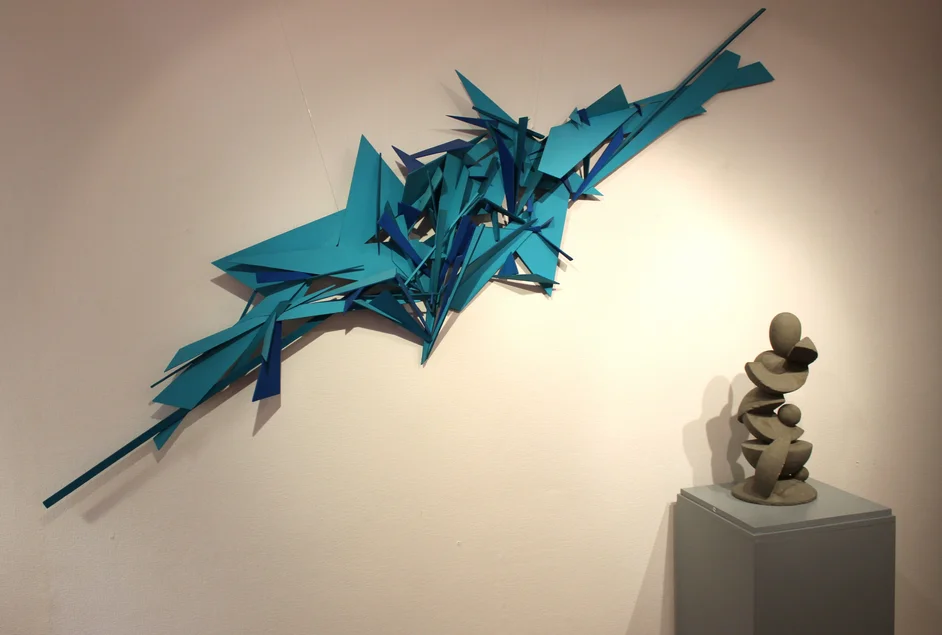 Ausstellungswand mit blau bemaltem, abstraktem Holz-Kunstwerk, rechts davor auf einem Sockel eine abstrakte Beton-Skulptur