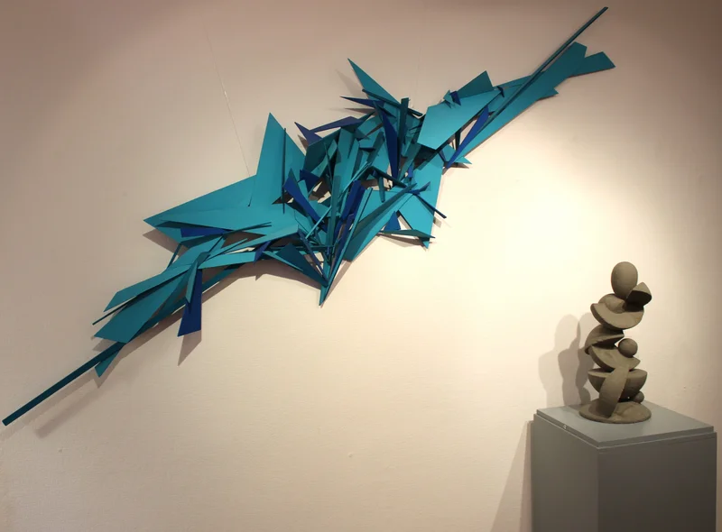 Ausstellungswand mit blau bemaltem, abstraktem Holz-Kunstwerk, rechts davor auf einem Sockel eine abstrakte Beton-Skulptur