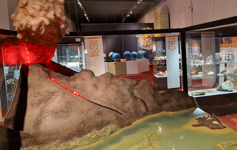Der Blick in die Ausstellung umfasst mehrere große Vitrinen. Die Vitrine im Vordergrund enthält das Modell eines Vulkanausbruchs, im Hintergrund sind Vitrinen mit Planetenmodellen und Fossilien zu sehen.
