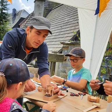 An einem im Freien stehenden Tisch hilft ein Erwachsener zwei Kindern beim Basteln von Miniatur-Wasserrädern.