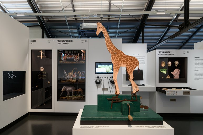 Die Welt des Puppentheaters – interaktiv erleben Ausstellungsraum der Basis-Ausstellung der Puppentheatersammlung Dresden im Kraftwerk Mitte: Im Vordergrund steht eine auffällige Figur: Die Giraffe ist das Modell einer Theatrum-mundi-Figur. Die Nachbildung macht die Bewegungsmechanik dieser historischen Theaterform anschaulich und erlebbar. Im Hintergrund sind mehrere Vitrinenschränke zu sehen, in denen Puppen verschiedener Größe und aus unterschiedlichen Materialien präsentiert werden.