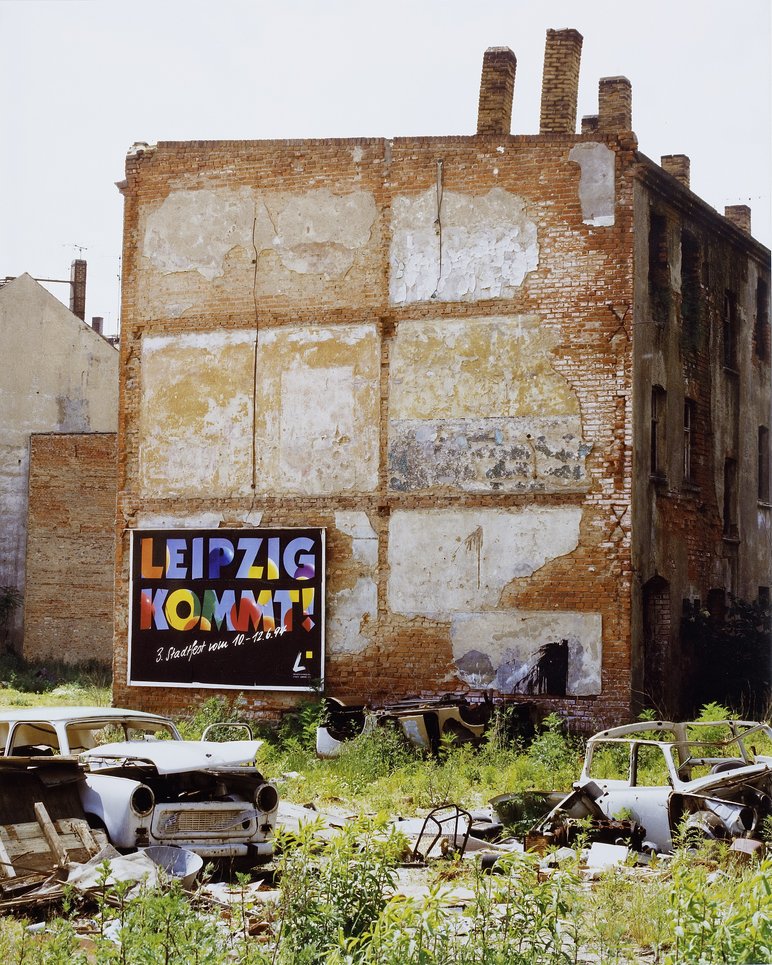 Auf der Grünfläche vor der abbröckelnden Stirnseite eines alten Ziegelgebäudes stehen die Überreste zweier DDR-Autos der Marke "Trabant", am Gebäude selbst hängt ein großes Werbeplakat mit der bunten Aufschrift "Leipzig kommt".