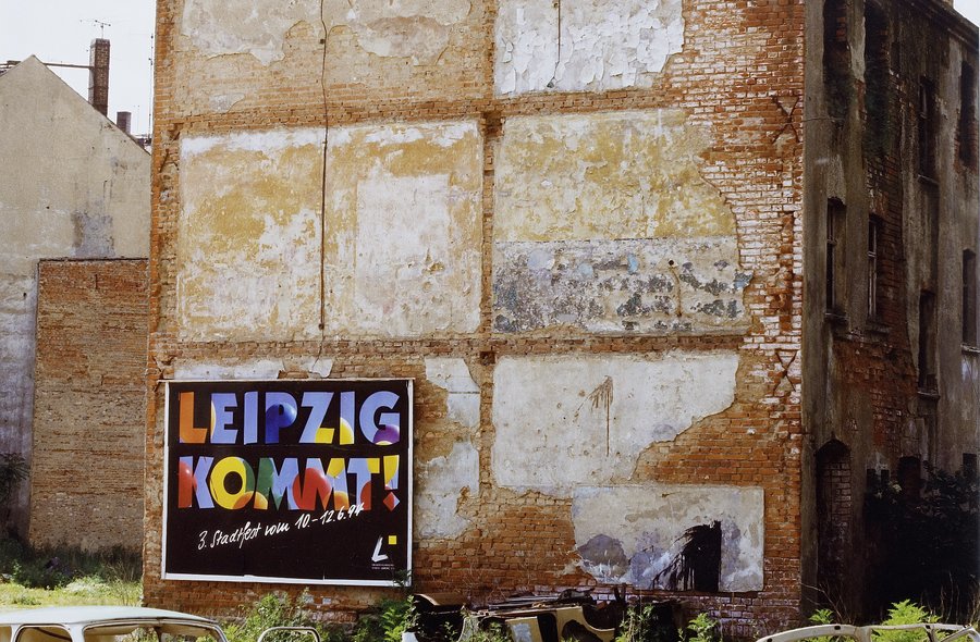Auf der Grünfläche vor der abbröckelnden Stirnseite eines alten Ziegelgebäudes stehen die Überreste zweier DDR-Autos der Marke "Trabant", am Gebäude selbst hängt ein großes Werbeplakat mit der bunten Aufschrift "Leipzig kommt".