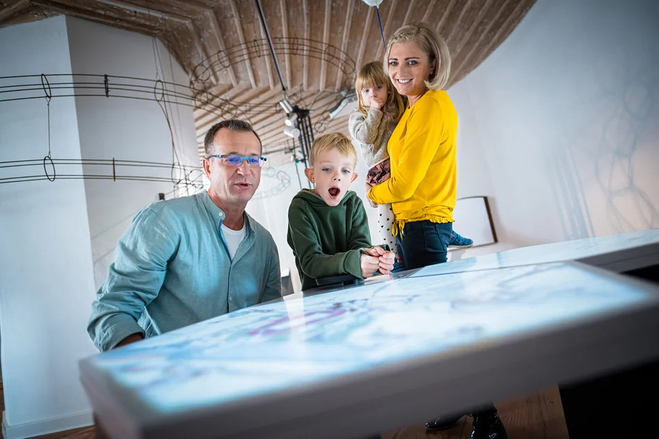 Eine Familie beugt sich über ein interaktives Element, im Hintergrund ist eine XXL-Kugelbahn zu sehen.