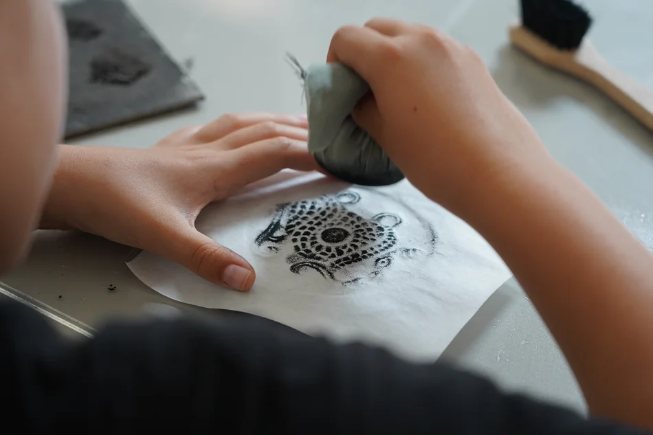 Ein Blatt Papier liegt auf einer mit einem Motiv versehenen Steinplatte. Darüber hält eine Hand einen Stein, der mit einem in schwarzer Tusche getränkten Stück Stoff umwickelt ist.