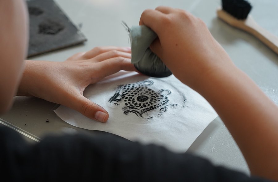 Ein Blatt Papier liegt auf einer mit einem Motiv versehenen Steinplatte. Darüber hält eine Hand einen Stein, der mit einem in schwarzer Tusche getränkten Stück Stoff umwickelt ist.