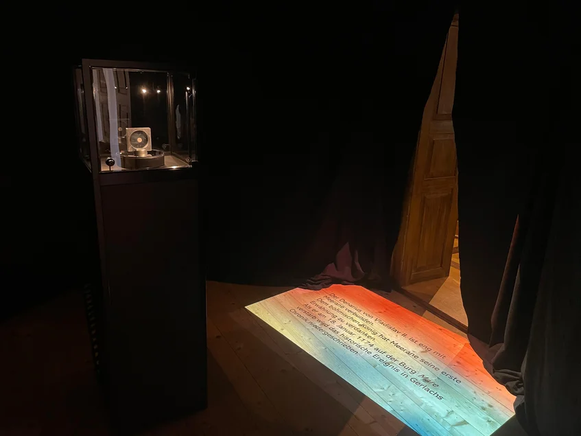 ln einem abgedunkelten Ausstellungsraum steht eine kleine Vitrine, in der sich die kleine Münze von Vladislav dem Zweiten befindet. Auf dem Boden davor ist die Projektion eines Ausstellungstextes erkennbar.