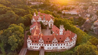 Von einer Drohne aufgenommenes Foto des historischen Schlosskomplexes im sommerlichen Abendlicht, umringt von einer Fülle von Laubbäumen