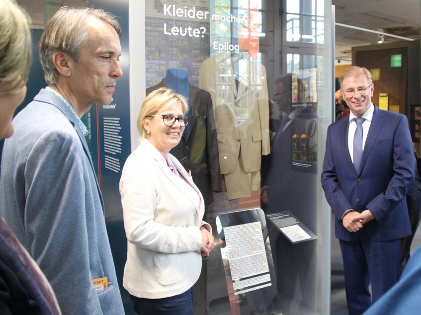 Eine Frau und zwei zu ihrer Linken und zu ihrer Rechten stehende Männer stehen vor einer großen Vitrine, in der Textilien präsentiert werden.