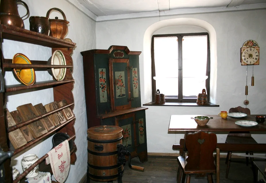 Bauernküche In einem weißgetünchten, historisch anmutenden Raum befinden sich Bauernmöbel aus dunklem Holz: ein offenes Regal mit historischen Küchenutensilien, ein bemalter Schrank, ein Esstisch mit Stühlen. Neben dem Fenster hängt eine mit Blumenmotiv verzierte Wanduhr.