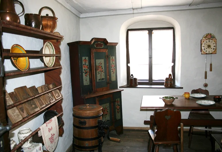 In einem weißgetünchten, historisch anmutenden Raum befinden sich Bauernmöbel aus dunklem Holz: ein offenes Regal mit historischen Küchenutensilien, ein bemalter Schrank, ein Esstisch mit Stühlen. Neben dem Fenster hängt eine mit Blumenmotiv verzierte Wanduhr.