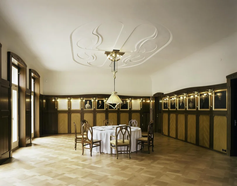 Das Foto zeigt das Esszimmer in der Villa Esche, entworfen von Henry van de Velde. In der Mitte steht ein runder Tisch mit sieben Stühlen, von der stuckverzierten Decke hängt eine elegante Lampe, an den holzvertäfelten Wänden hängen mehrere gemalte Porträts.
