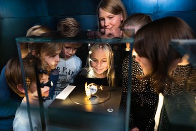 Im Dunkeöln blicken eine Museumsmitarbeiterin und eine kleine Kindergruppe in eine mit Taschenlampen angestrahlte Vitrine, in der eine historische Taschenuhr präsentiert wird.