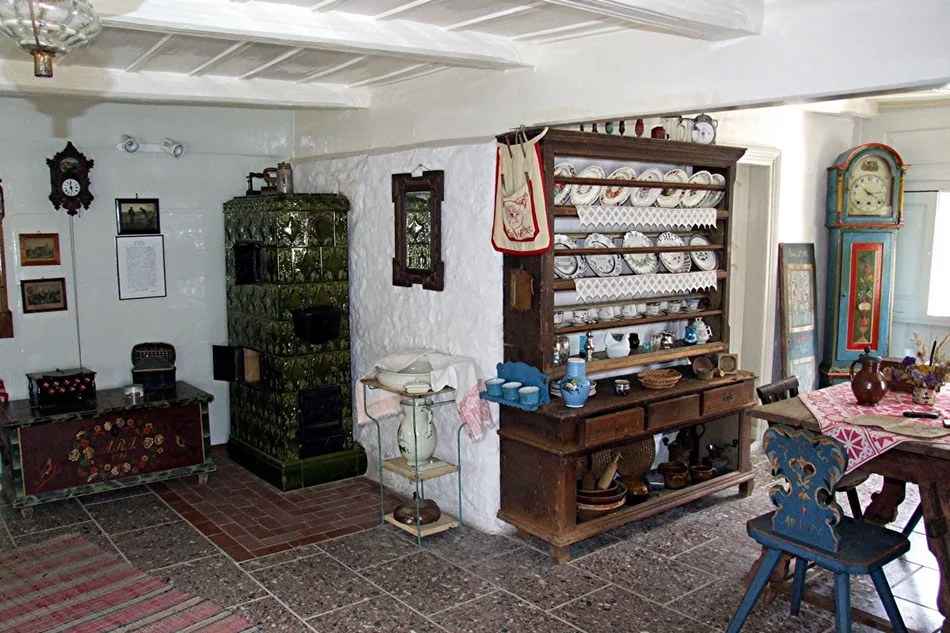 Blick in die Heimatstube Historische Bauernstube mit traditioneller Einrichtung, darunter ein grüner Kachelofen, eine verzierte Truhe, ein rustikaler Geschirrschrank mit Porzellan, eine große Standuhr und ein Esstisch mit bestickter Tischdecke und Holzstühlen