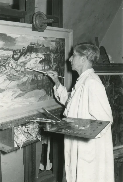 Schwarzweiß-Foto einer kurzhaarigen Frau mittleren Alters, die malend vor einer Staffelei steht