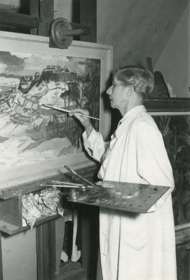 Schwarzweiß-Foto einer kurzhaarigen Frau mittleren Alters, die malend vor einer Staffelei steht