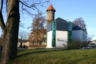 Historisches Gebäude mit einem modern wirkenden Anbau, auf dem "Museum" steht", dahinter ragt ein alter Turm empor.