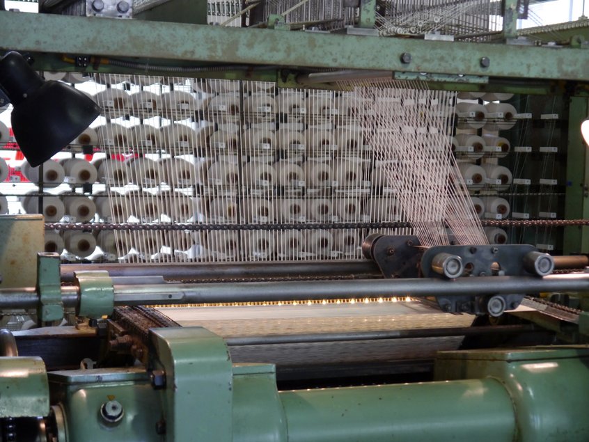 Das Foto zeigt eine Malimo-Maschine aus DDR-Zeiten im Betrieb. Verarbeitet werden im Bildbeispiel dutzende Spulen weißen Garns.