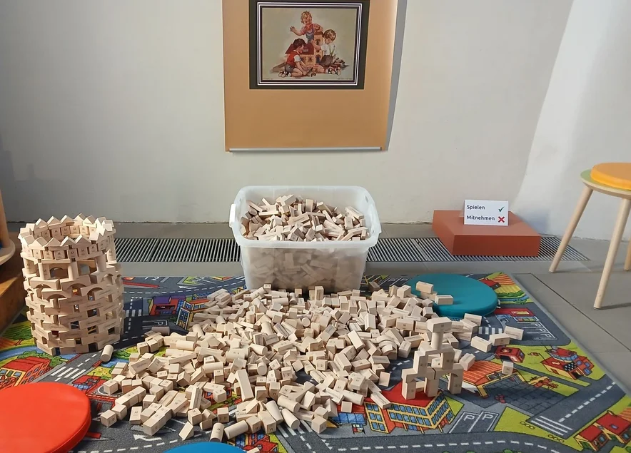 Auf einem Spielteppich liegt ein großer Haufen Holzbausteine, daneben steht ein aus Holzbausteinen errichteter Turm.