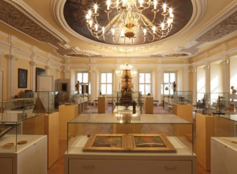 Foto eines prächtigen Barocksaals, in dem sich eine Vielzahl von Vitrinen mit unterschiedlichen Exponaten befinden und von dessen bemalter und reich verzierter Decke ein großer Kronleuchter hängt