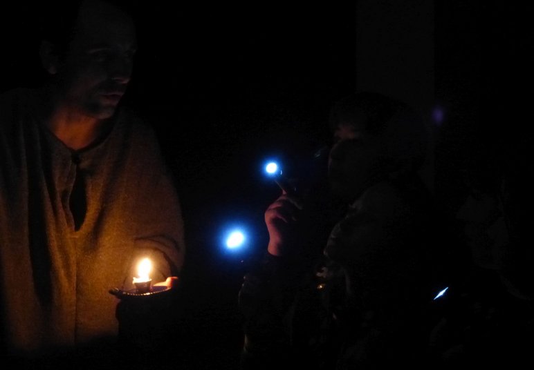 DAs Foto zeigt zwei Kinder im Dunkeln mit Taschenlampen sowie einen Mann in historischem Kostüm mit einem Kerzenhalter in der Hand.