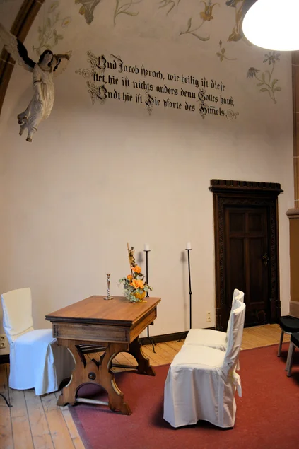 Ein Tisch mit Stühlen für einen Standesbeamten und ein Brautpaar, an der mit Blumen verzierten Wand die Figur eines Engels