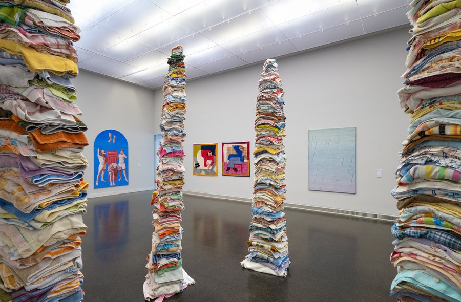 In einem Ausstellungssaal stehen vier aus verschiedenfarbenen, aufeinandergestapelten Textilien bestehende Stelen. An zwei Ausstellungswänden im Hintergrund hängen weitere Kunstwerke.