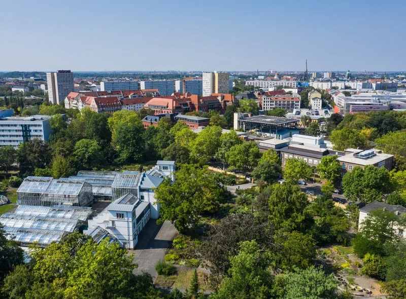 Luftaufnahme über Gewächshäuser und viel Grün, im Hintergrund urbane Bebauung der Stadt Leipzig