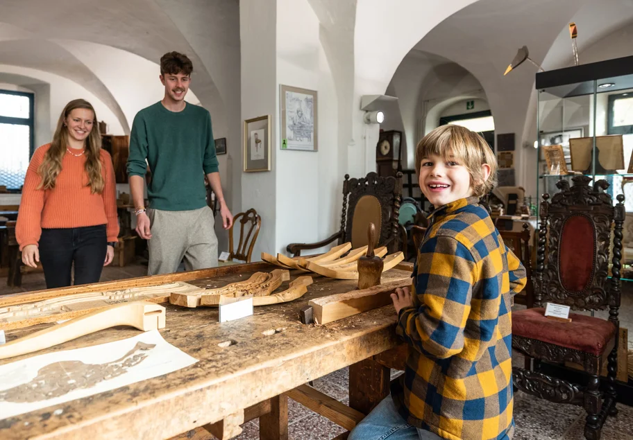 Drei Personen stehen um eine Werkbank, im Hintergrund historische Stühle