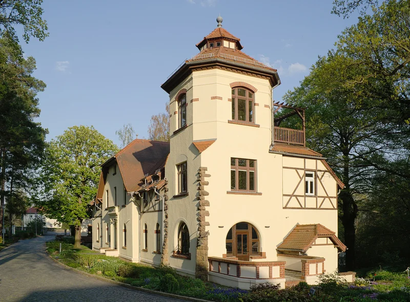 Helles historisches Haus mit Turm, Fachwerkdetails, umgeben von Bäumen und Weg