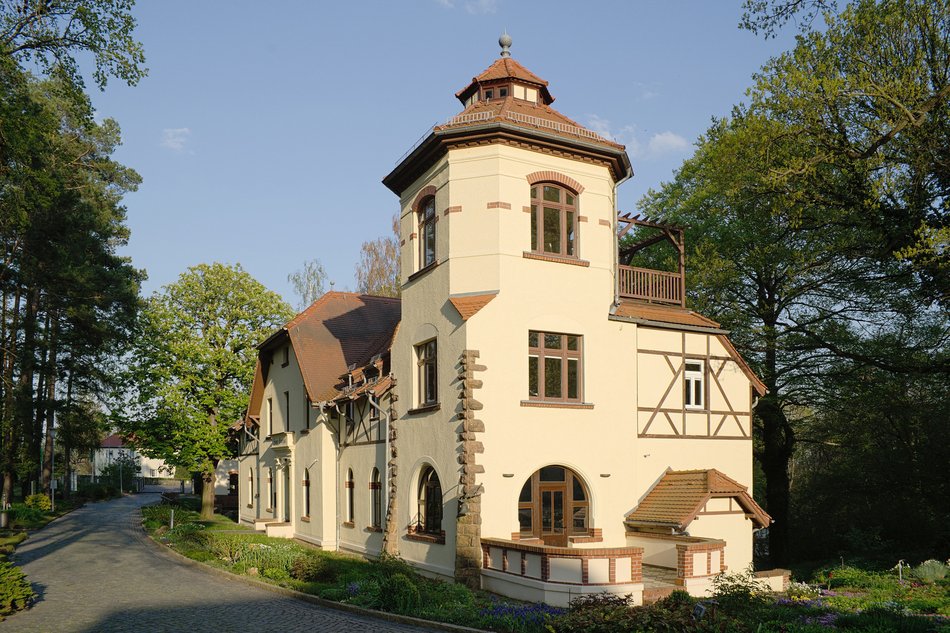 Helles historisches Haus mit Turm, Fachwerkdetails, umgeben von Bäumen und Weg