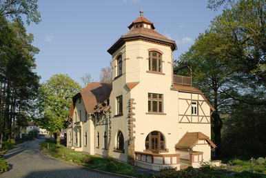 Haus Energie – das ehemalige Wohnhaus Wilhelm Ostwalds Helles historisches Haus mit Turm, Fachwerkdetails, umgeben von Bäumen und Weg