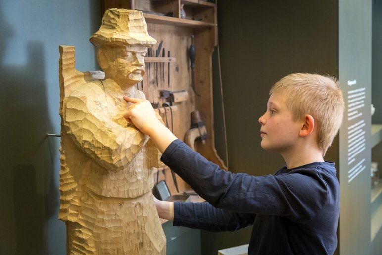In einem Ausstellungsraum berührt ein Junge die aus Holz geschnitzte, nahezu lebensgroße Figur eines Jägers.