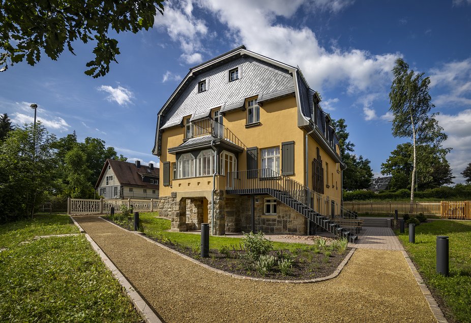 Sommerliche Außenansicht einer Villa aus dem frühen 20. Jahrhundert mit sandfarbener Fassade und schiefergedecktem Dachfirst. Seitlich führt eine angebaute Treppe zu einem Erker im Hochparterre.