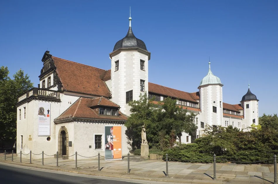 Sommerliche Außenansicht eines Schlösschen-artigen historischen Gebäudes mit weißen Außenwänden und drei regelmäßig entlang der Längsseite angeordneten Türmchen